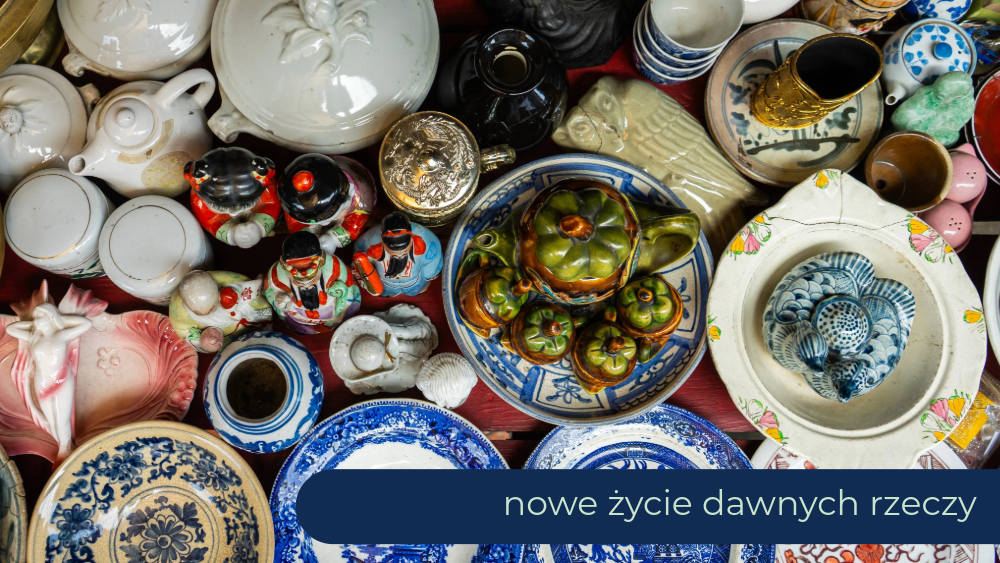 Widok z góry na dużą kolekcję różnorodnej, antycznej porcelany i ceramiki, w tym talerze, dzbanki i figurki. W centrum kompozycji znajduje się zielono-brązowy ceramiczny czajnik i filiżanki w stylu ludowym, otoczony niebiesko-białymi oraz jednolitymi białymi elementami. Na dole obrazu widoczny jest granatowy pasek z napisem "nowe życie dawnych rzeczy".